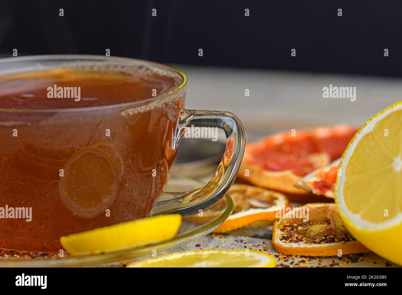 Cup with hot tea and steam on black. Glass cup of black tea with cinnamon sticks, anise star, lemon and dried fruit on white wooden table background. Macro image Stock Photo