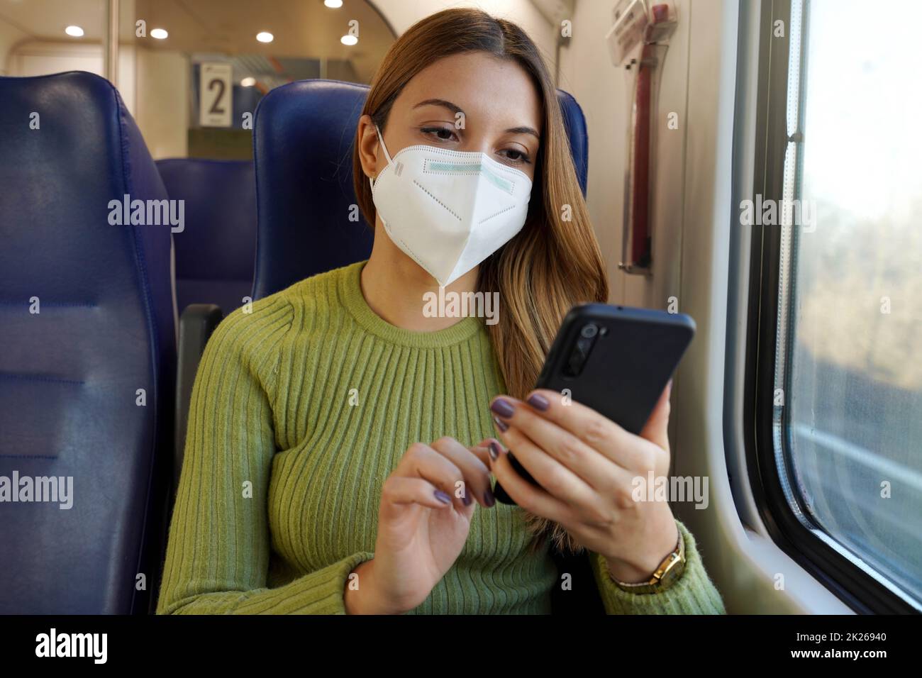 Relaxed woman on train wearing medical face mask using smart phone app. Commuter with protective mask traveling sitting in business class texting on mobile phone. Travel safely on public transport. Stock Photo