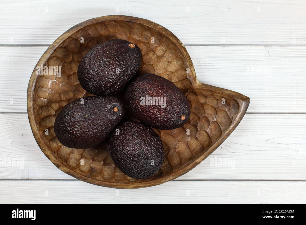 Four whole ripe brown avocados in wooden carved bowl on white boards ...