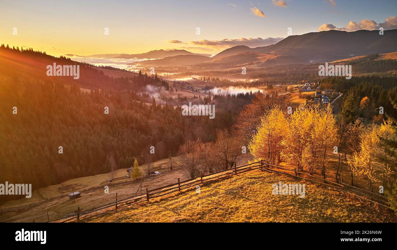 Colorful yellow trees on hill. Autumn sunrise, foggy woodland. Forest covered by low clouds. Fall rural landscape. Village in misty valley. Stock Photo