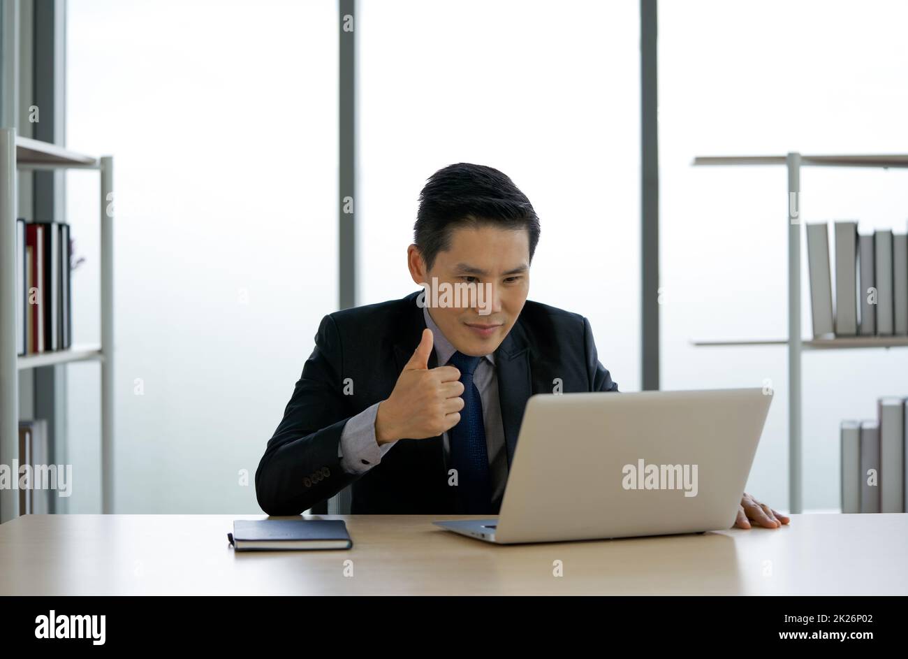 Asian businessman in black suit raise finger thumb up via video conference on computer monitor. Concept of teleconferencing through wireless communication. Stock Photo