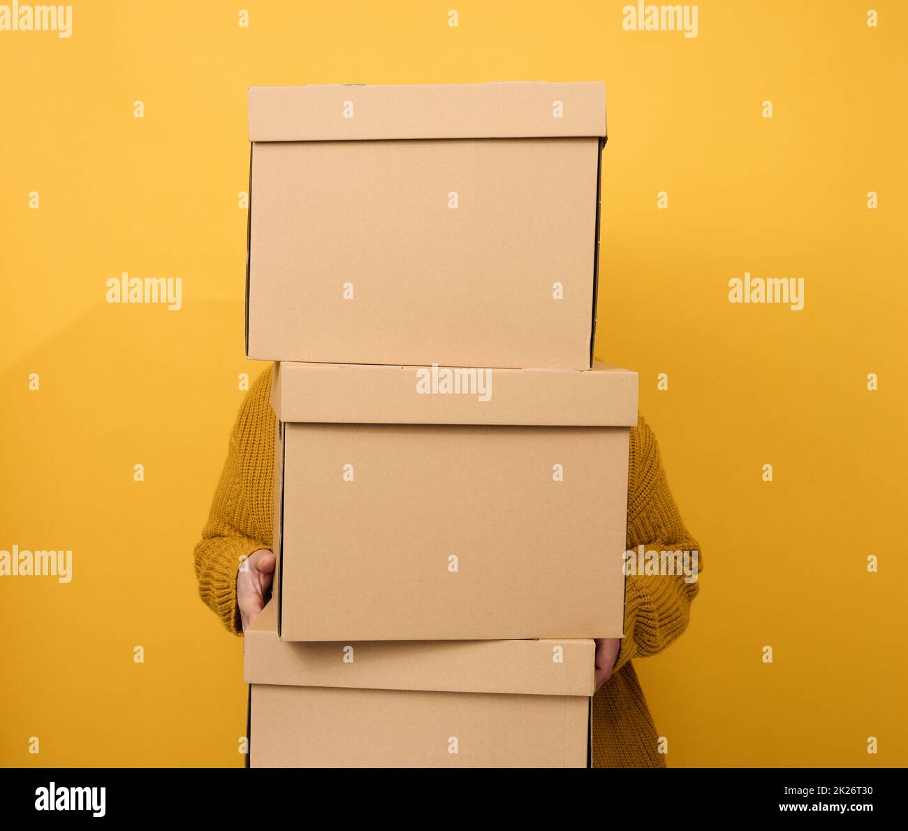 a woman in a sweater holds a stack of brown cardboard boxes on a yellow background. Relocation Stock Photo