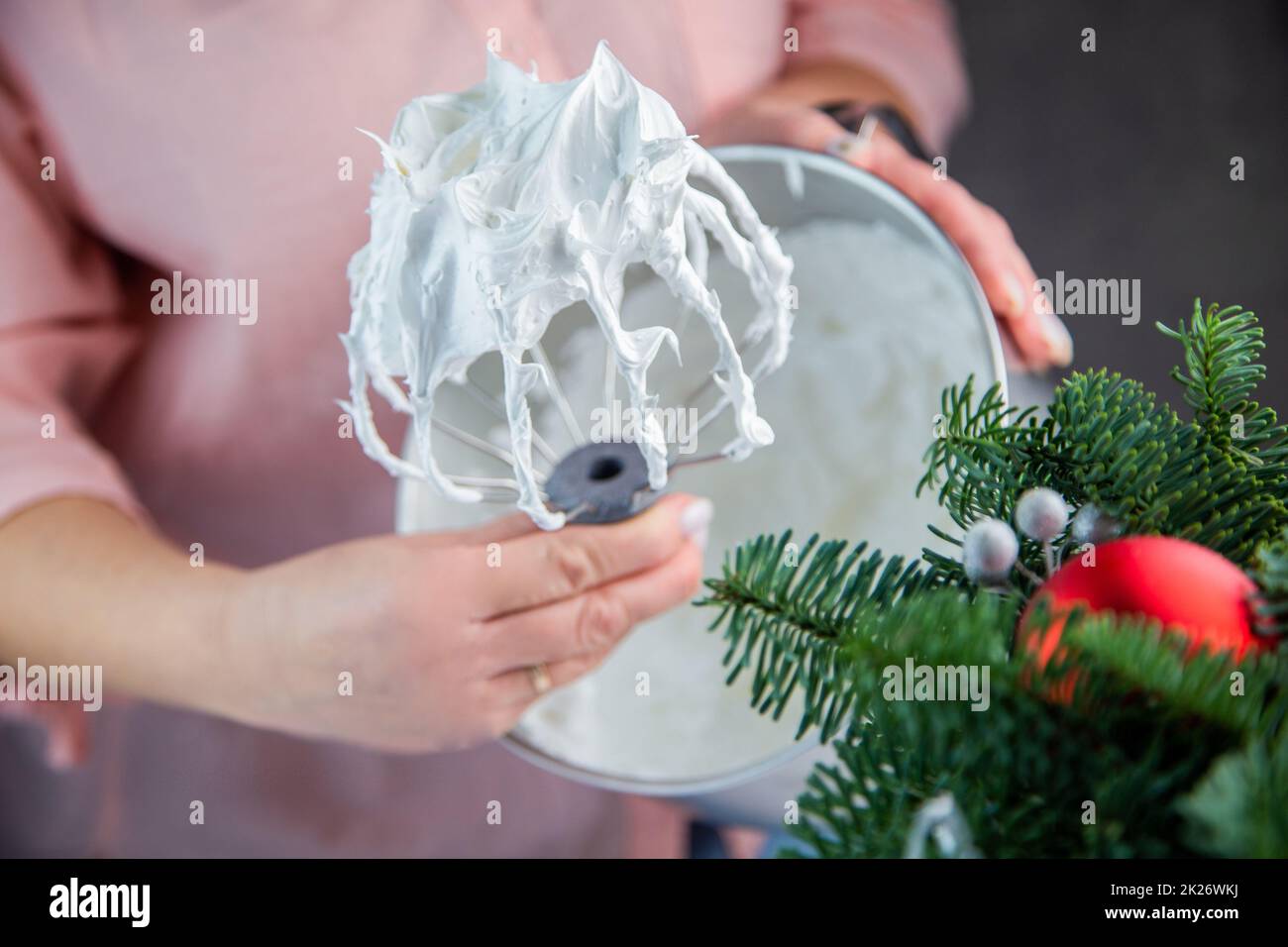 The whisk of an electric mixer in a white mass of meringue with standing peaks is held in the hand of a pastry chef against the background of an iron bowl, the branches of a Christmas tree and New Year's toys are visible from the side. Stock Photo