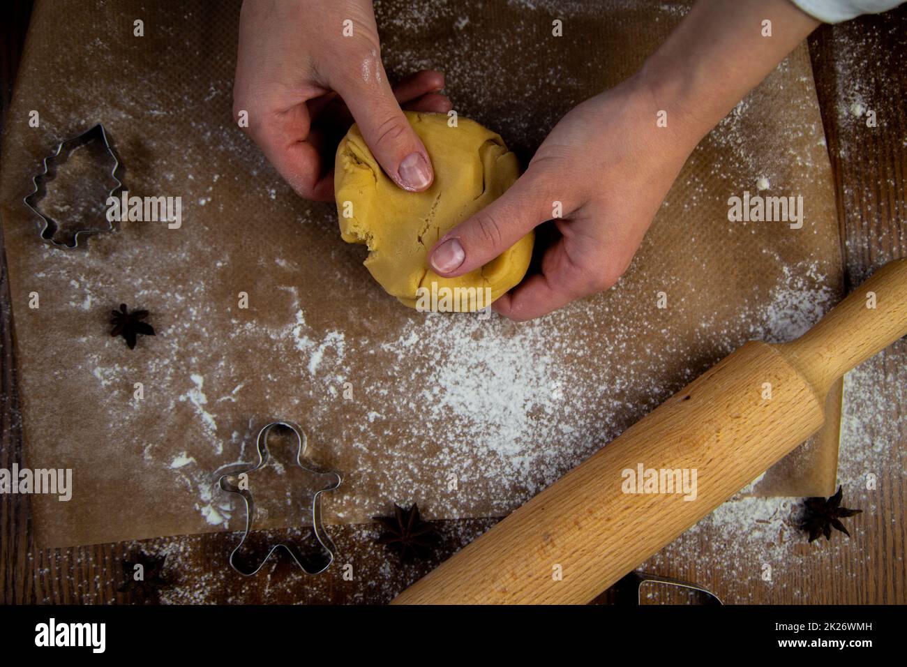 In the foreground is a table with baking parchment lying on it, on which there is dough that the cook's hands are crumpling, a rolling pin, baking molds and flour are scattered around. Stock Photo