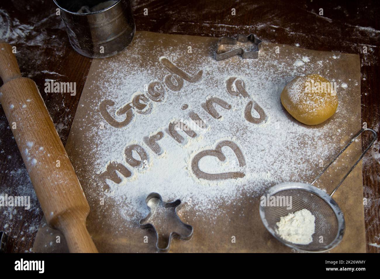 On the baking paper on the flour is written Good morning and heart, against the background of dough, rolling pins and cookie cutters Stock Photo