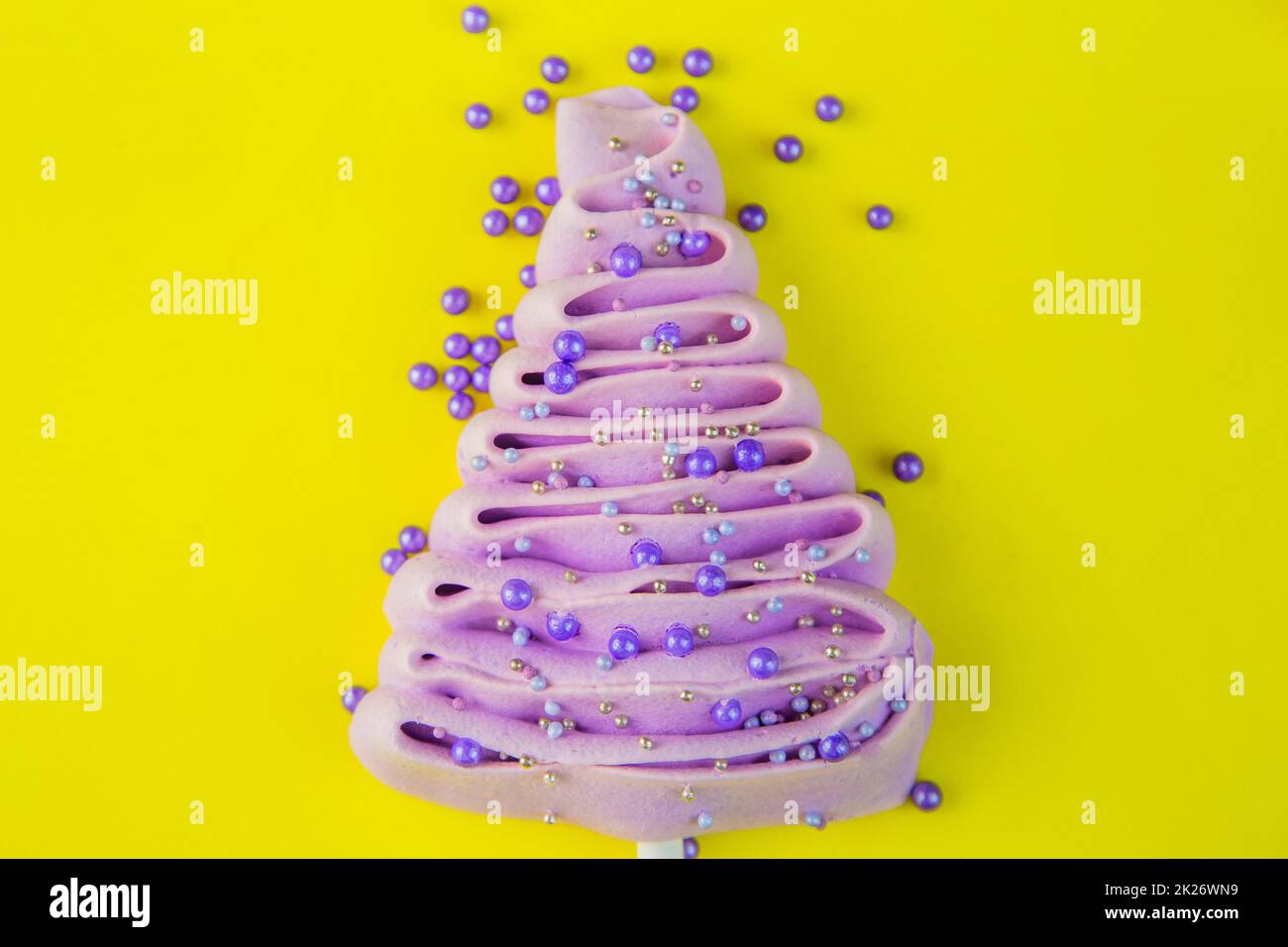 Pale purple Christmas tree made of meringue on a stick, decorated with purple and silver balls on a bright yellow background, purple powder balls are scattered near the tree. Stock Photo