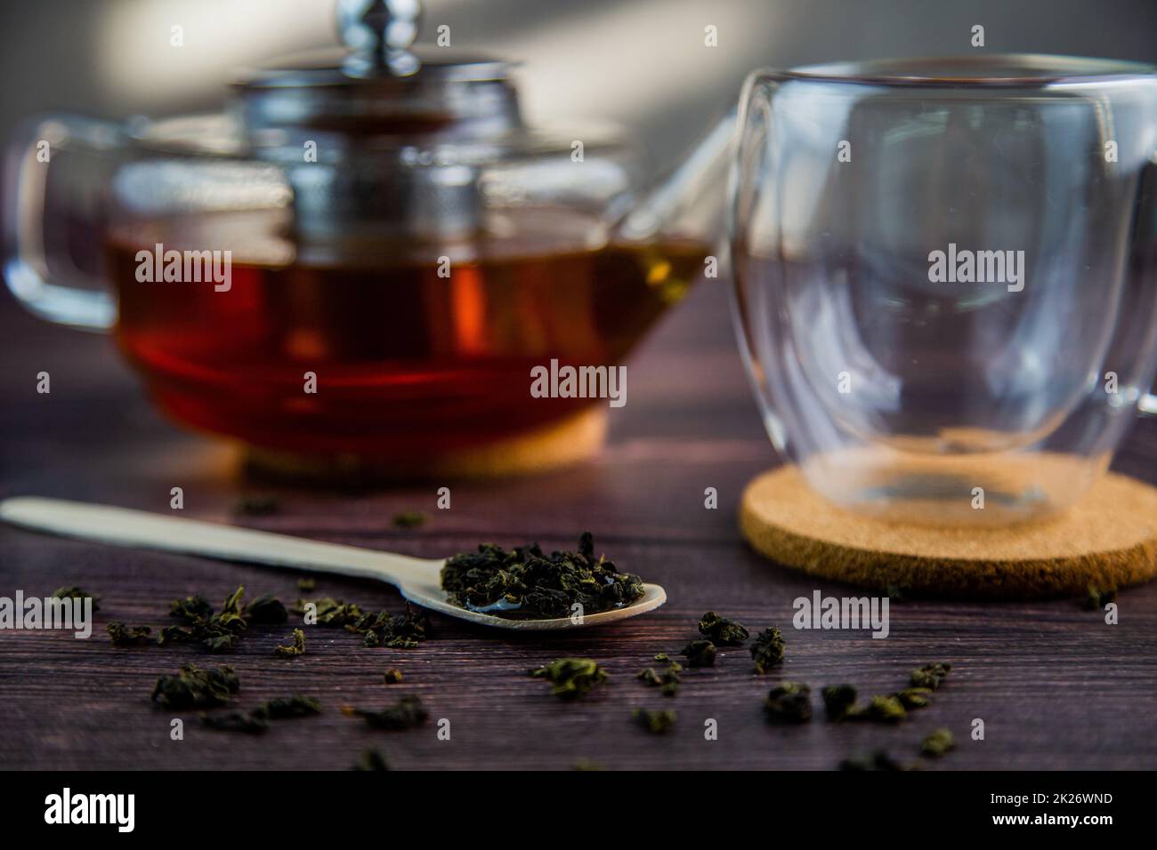 A wooden spoon with tea lies against a dark wood background, a mug and a teapot are visible from behind, a photo in dark colors. Stock Photo