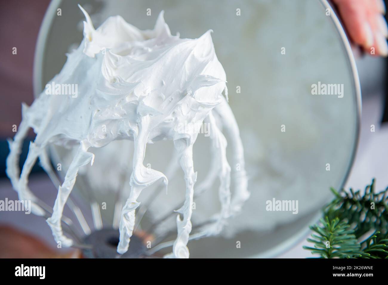 A whisk from a metal mixer in a white mass of bizet on the background of a bowl, the hands of a pastry chef and the branches of a Christmas tree are visible Stock Photo