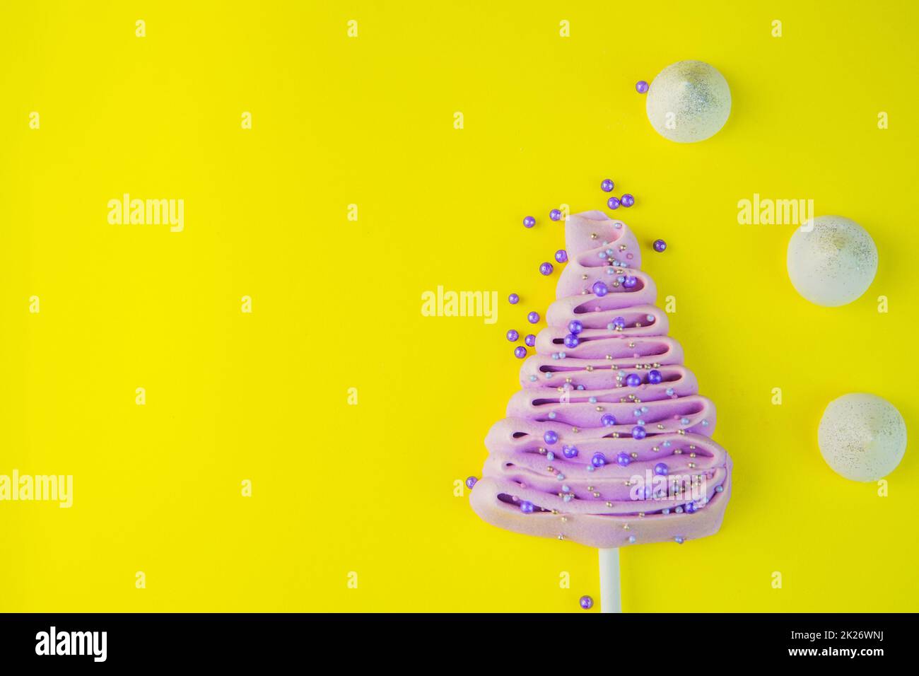 Pale purple Christmas tree made of meringue on a stick, decorated with purple and silver balls on a bright yellow background, purple powder balls and white snowdrifts are scattered near the tree. Stock Photo