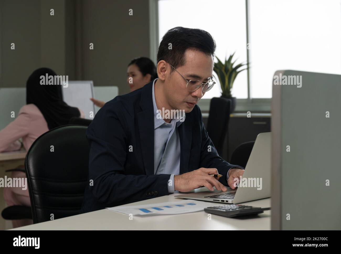Businessman working on laptop computer sitting at office desk in her workstation with colleagues in background. Stock Photo