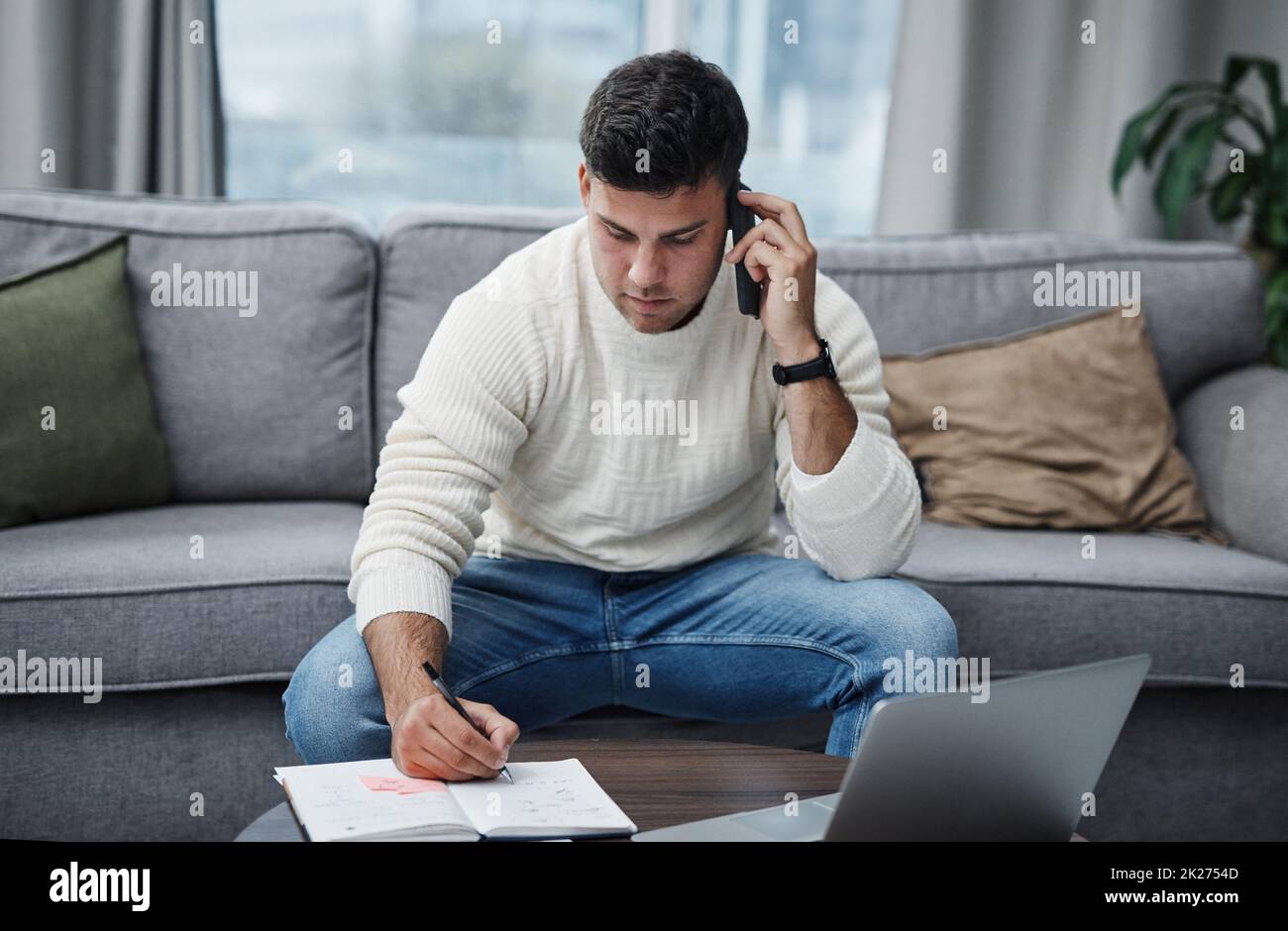 Part living space, part work space. Shot of a young man using a laptop and smartphone while writing in a notebook at home. Stock Photo