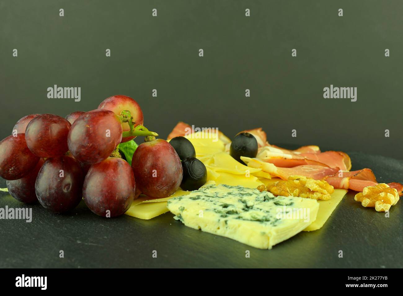 Assorted cheeses on black slate board. Brie cheese, cheddar slices, walnuts, grapes, olives and