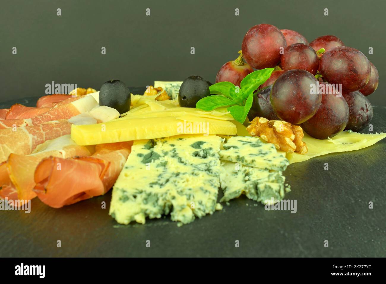 Assorted cheeses on black slate board. Brie cheese, cheddar slices, walnuts, grapes, olives and