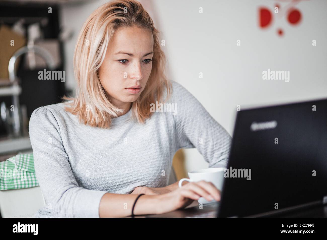 Female freelancer in her casual home clothing working remotly from her dining table in the morning. Home kitchen in the background. Stock Photo