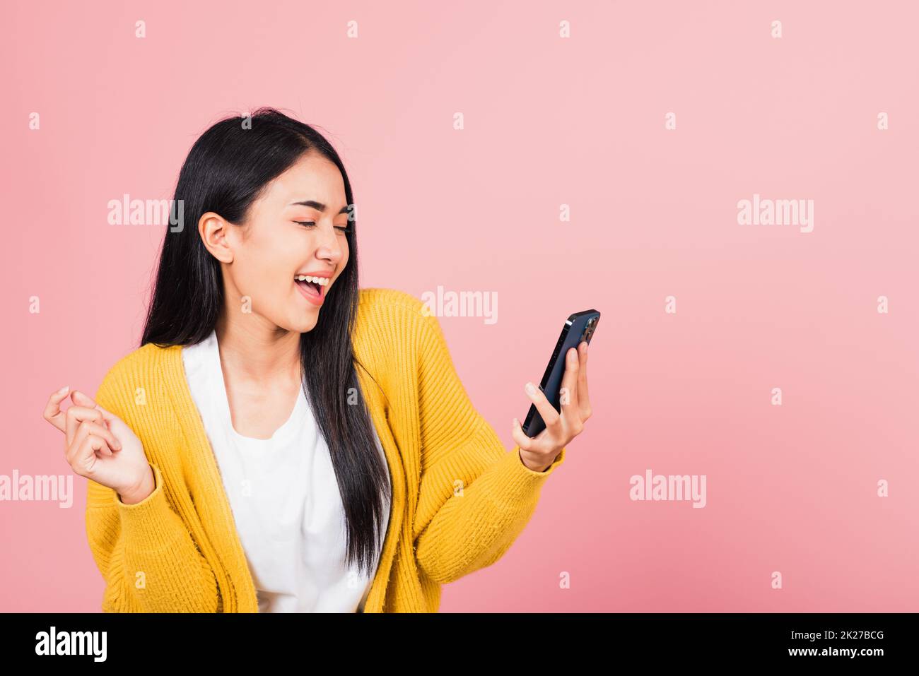 Happy Asian portrait beautiful cute young woman excited laughing holding mobile phone, studio shot isolated on pink background, female using funny smartphone making winner gesture Stock Photo