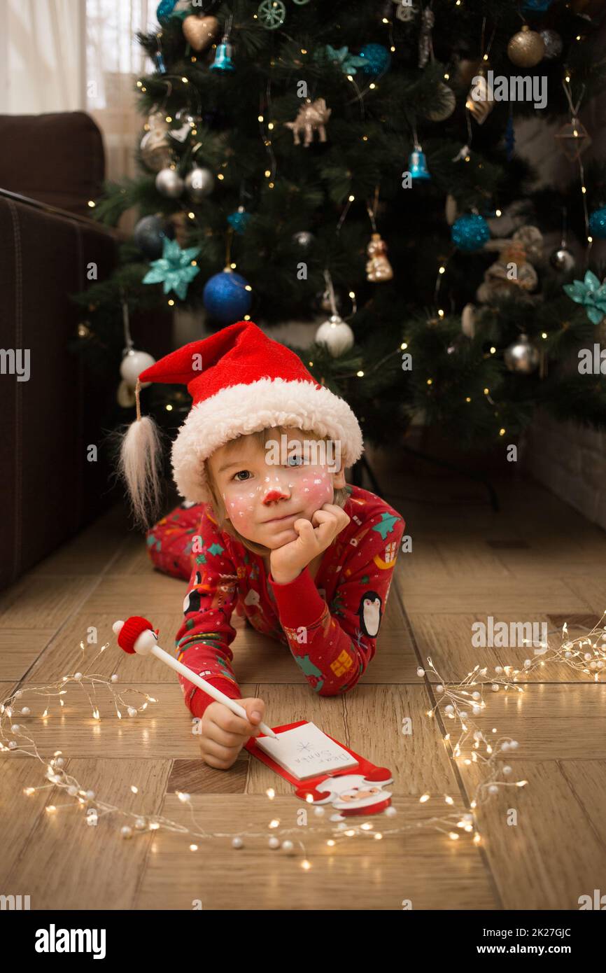 child in Christmas pajamas, Santa's hat and deer make-up writes letter to Santa Claus lying on floor in front of Christmas tree. winter holidays, inte Stock Photo