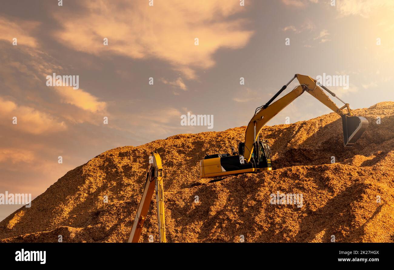 Backhoe working on huge sawdust pile in paper production factory. Bucket of digger digging wood chips. Pile of wood chips from pulp mill against the sky. Pulp and paper industry. Bulldozer  working. Stock Photo