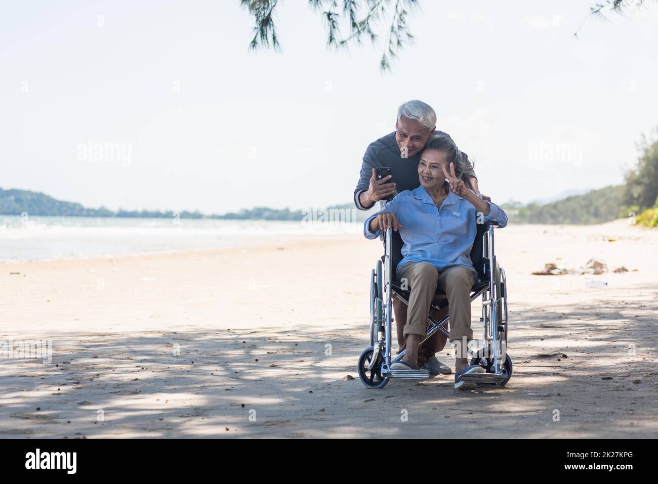 elderly woman sitting in wheelchair and husband is a wheelchair user smartphone taking selfie on the beach Stock Photo