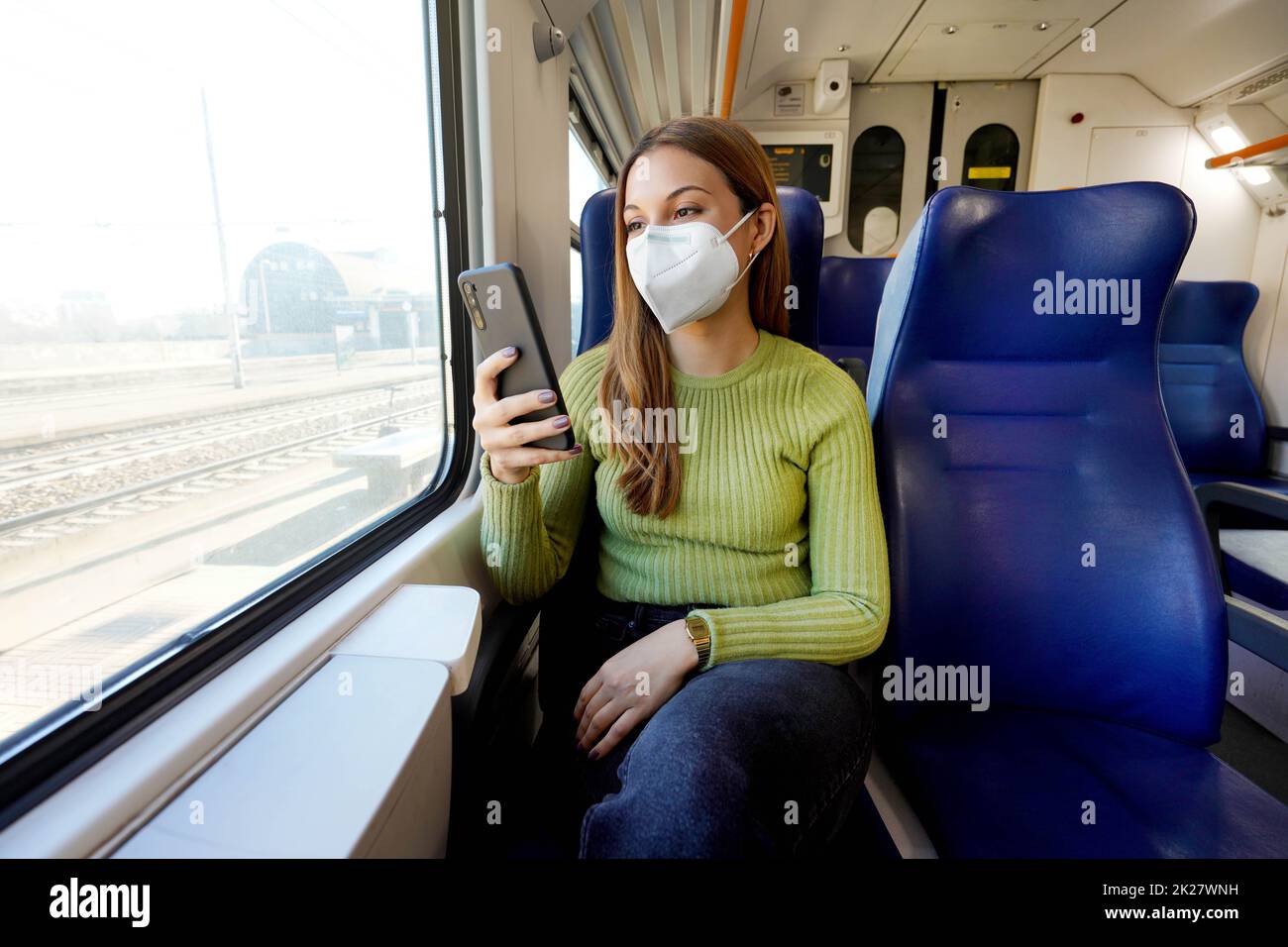 Young woman wearing medical mask relaxing in train seat while using smartphone app. Business woman enjoying view texting on mobile phone. Travel safety lifestyle. Stock Photo