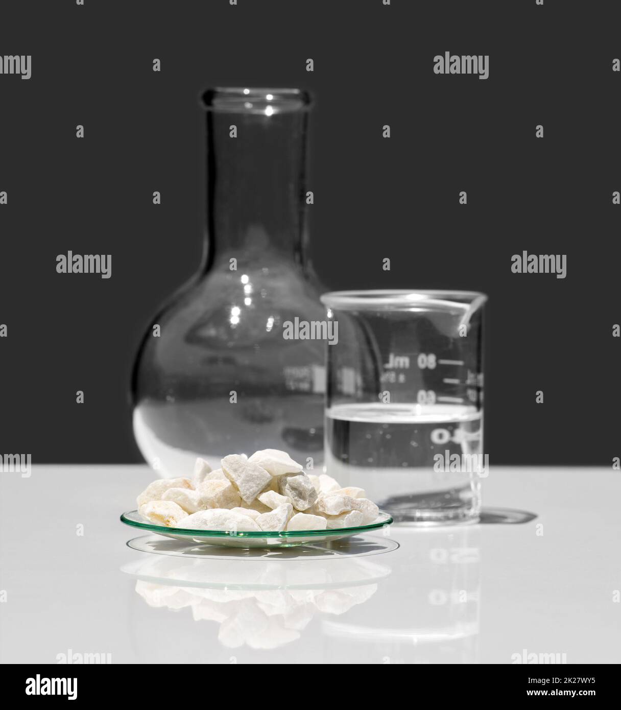Calcium carbonate Chip in Chemical Watch Glass place next to crystal clear liquid in Beaker and Flat Bottom Flask (Borosilicate Glass). Closeup chemical ingredient on white laboratory table. Stock Photo