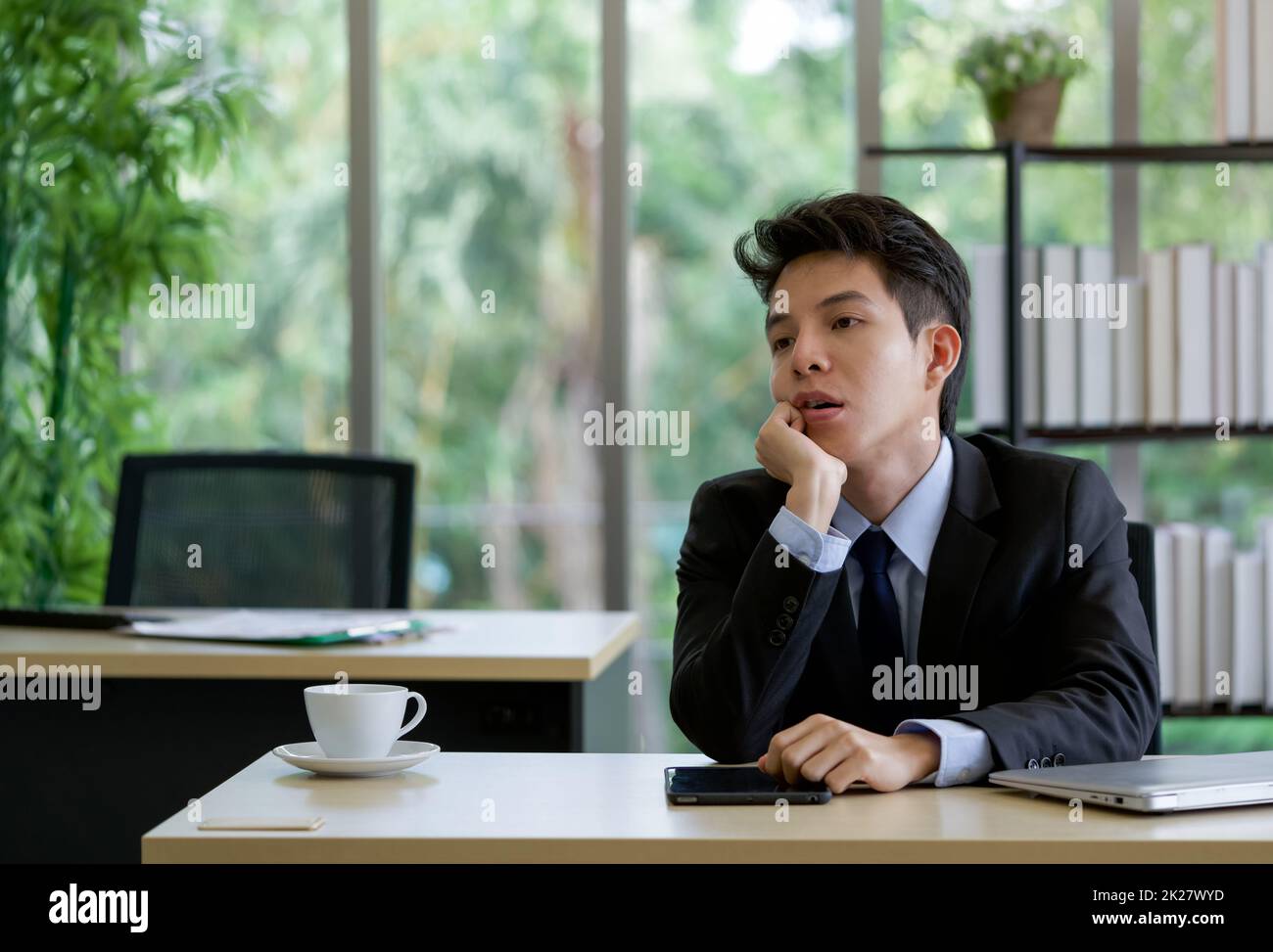 Young asian employee sit with absent-minded in the office on a table with tablet, laptop computer and a cup of coffee. Stock Photo