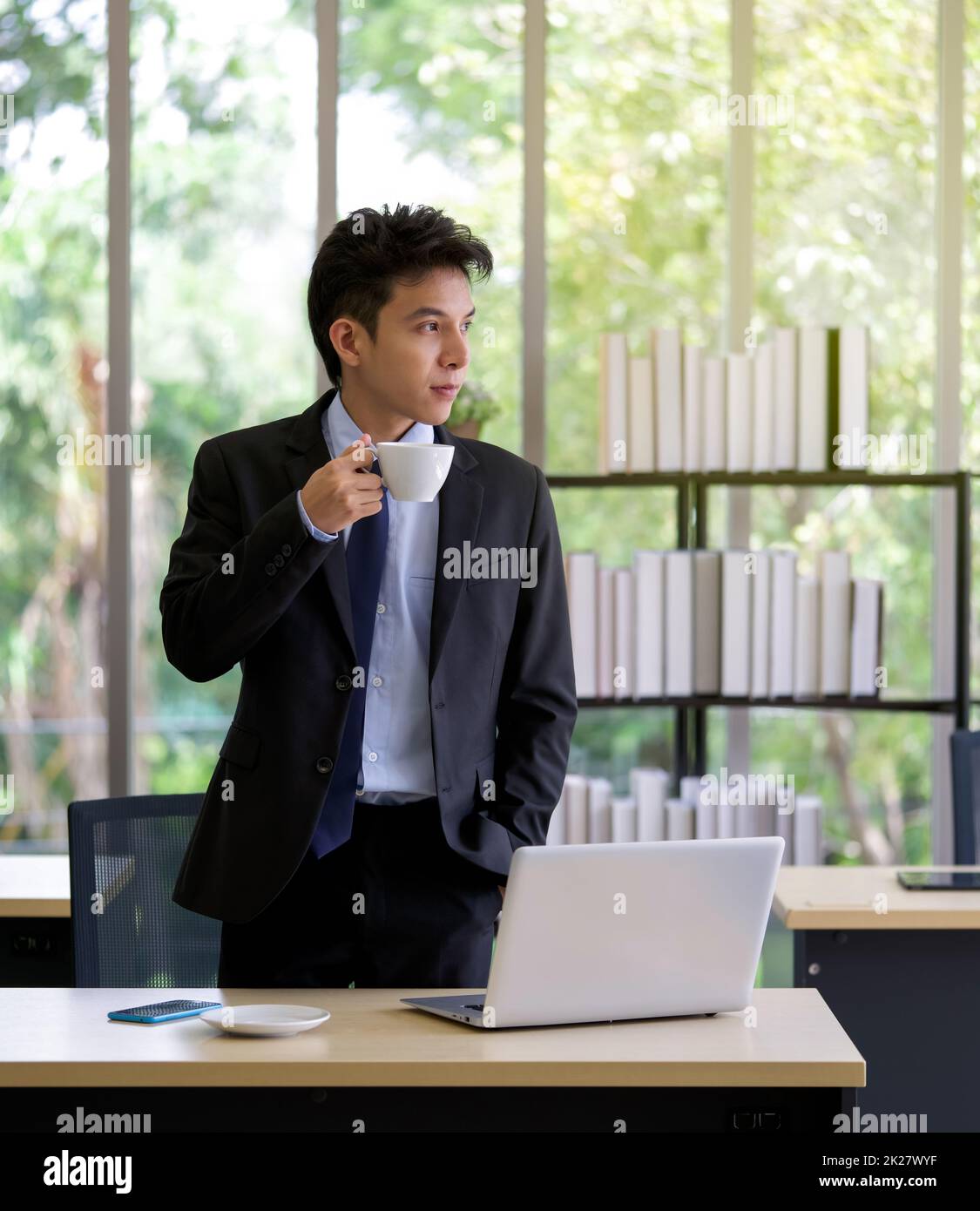 Young asian businessman in black suit drinking coffee while looked outside and thinking of something. Stock Photo