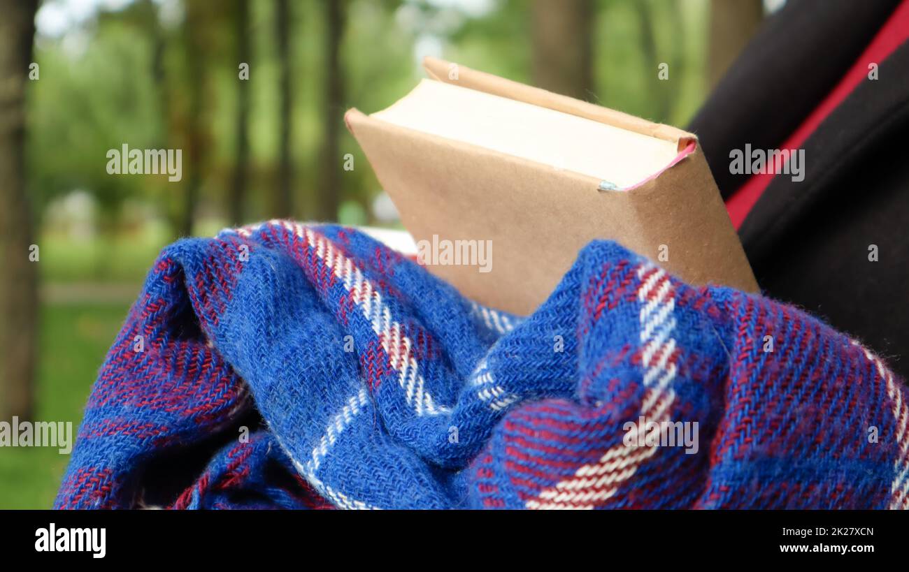A book with a blue checkered woolen blanket or plaid in the hands of a woman wearing a sweater and black coat in the park. Warm and sunny weather. Soft, cozy photograph. Close-up Stock Photo