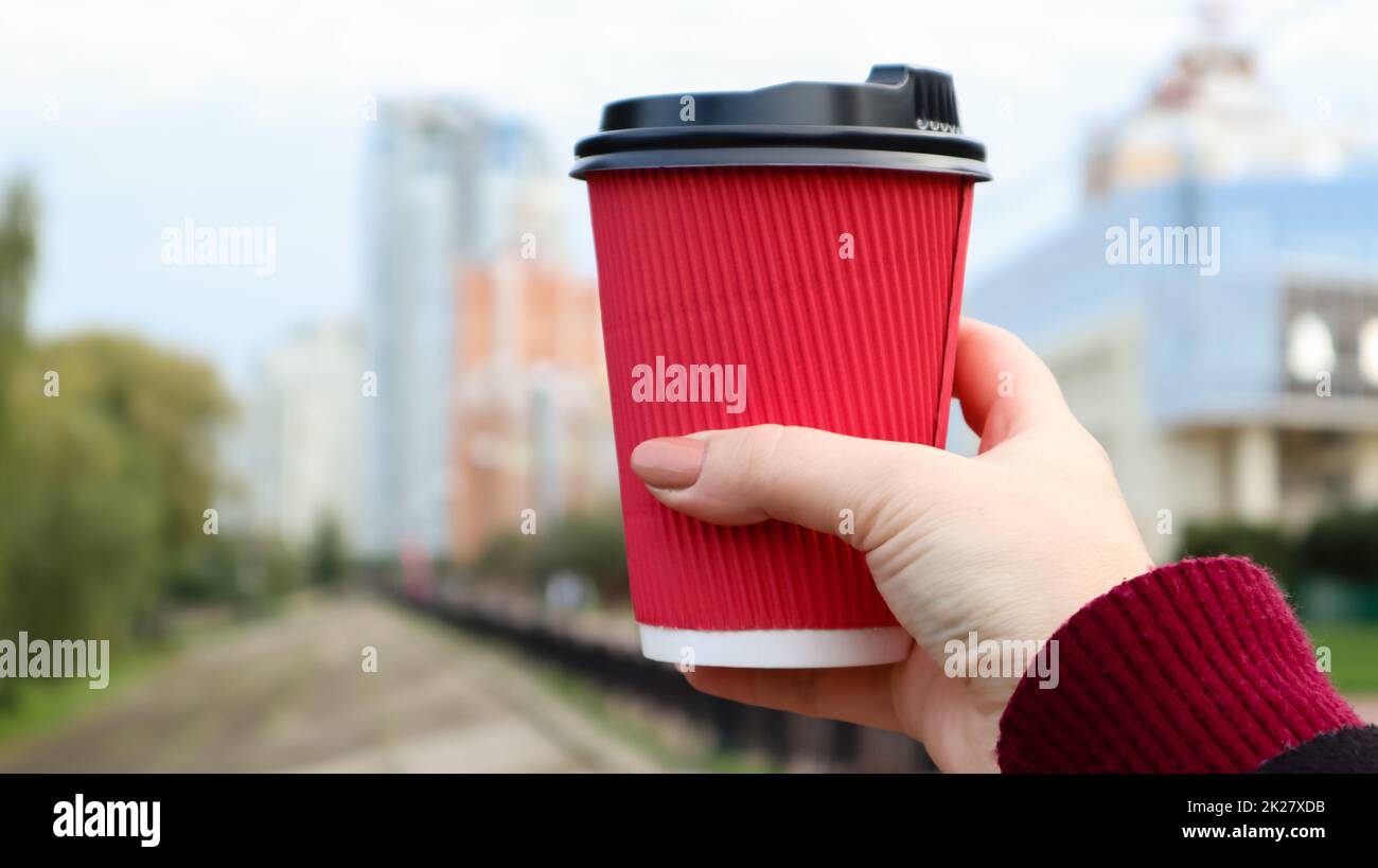 A young girl holds a red disposable paper cup with coffee or other hot drink in her hands during the cold season. Blurred street in the background. To go, away. No ecology. Stock Photo