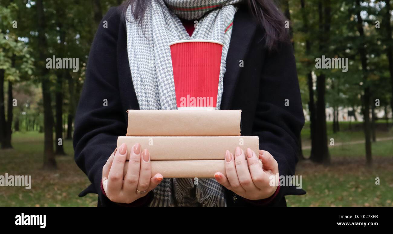A young woman in a black coat and a gray scarf holds a stack of books and a glass with coffee in her hands on the background of an autumn park. On open air. Education, close hands of the girl. Stock Photo