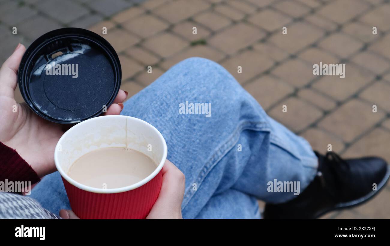 Female hands are holding morning takeaway coffee and closing a paper cup with a lid while sitting on a park bench, close-up of hands. Stock Photo