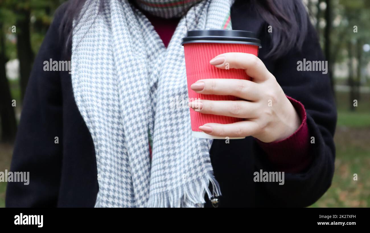 A young girl in a coat holds morning coffee with her while walking in the park. Hand holding paper cup of coffee in green park. Takeaway cappuccino. Close-up. Stock Photo