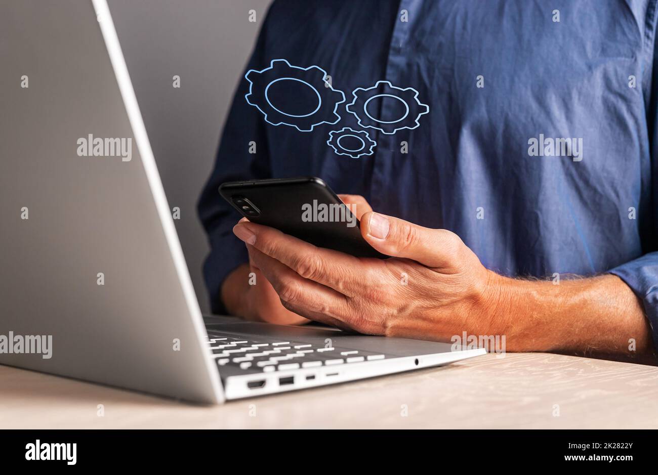 Man holding phone with virtual gears while working on laptop. Mobile settings or strategy, process organization, business idea concept. High quality photo Stock Photo