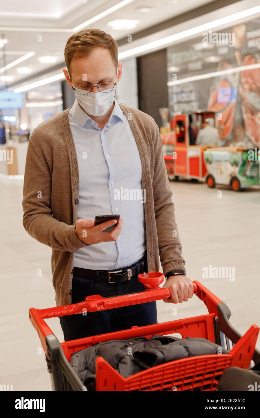 Male customer in medical face mask walking with shopping trolley in mall. Using mobile check list Stock Photo