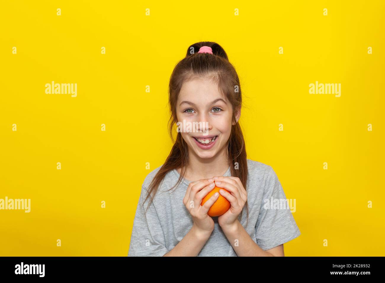 Little smiling cute girl in gray t-shirt holding fresh an orange over yellow background. Healthy lifestyle and clean eating concept Stock Photo