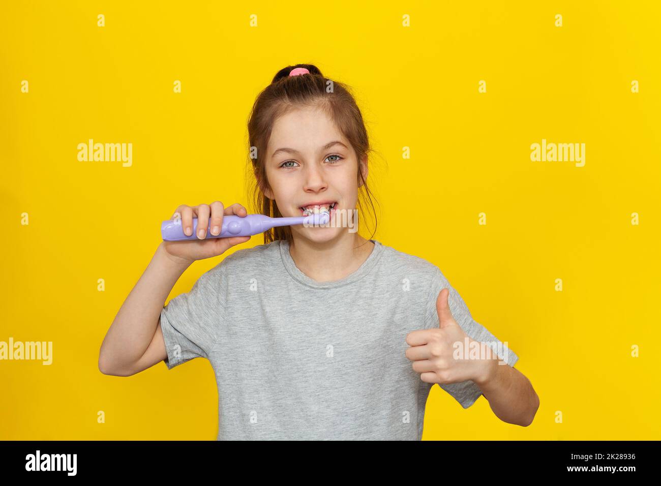 Beautiful brown-haired teenage girl holding brushing her teeth with an electric toothbrush on a yellow background, Children nutrition and oral hygiene concept Stock Photo