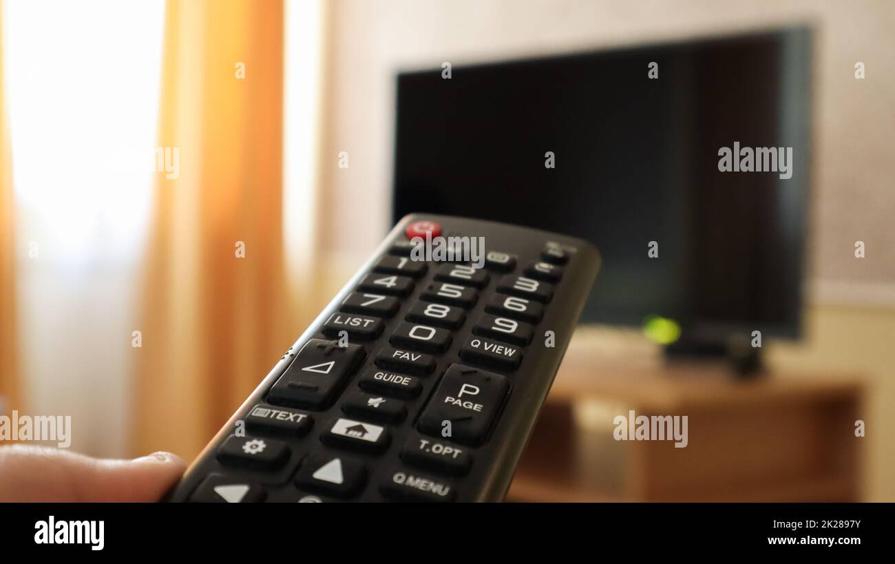Hand holding a television remote control and surfing programs on television. watch, turn on or off the TV in the living room or bedroom on the black-screen nightstand. Copy space. Stock Photo
