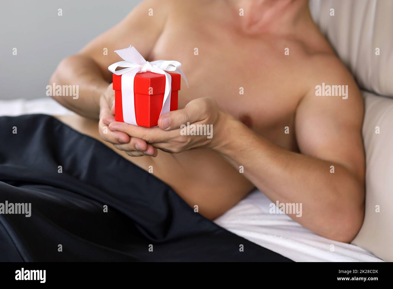 Muscular man lying in bed under a black blanket with red gift box in hands. Concept of birthday, Christmas holiday, New Year or Valentine's day Stock Photo