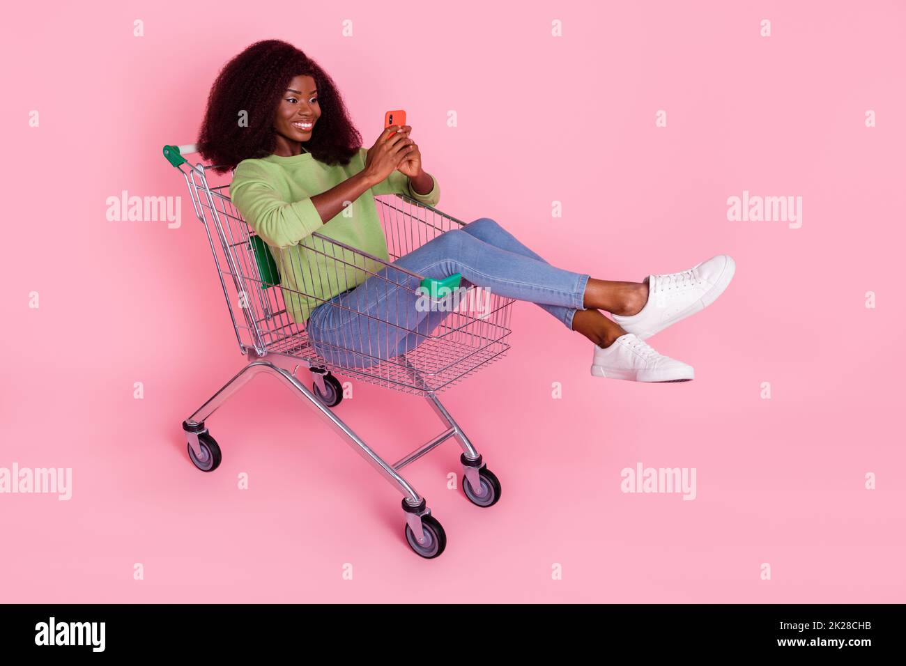 Full length photo of pretty cute lady wear green sweater sitting shopping tray chatting gadget isolated pink color background Stock Photo