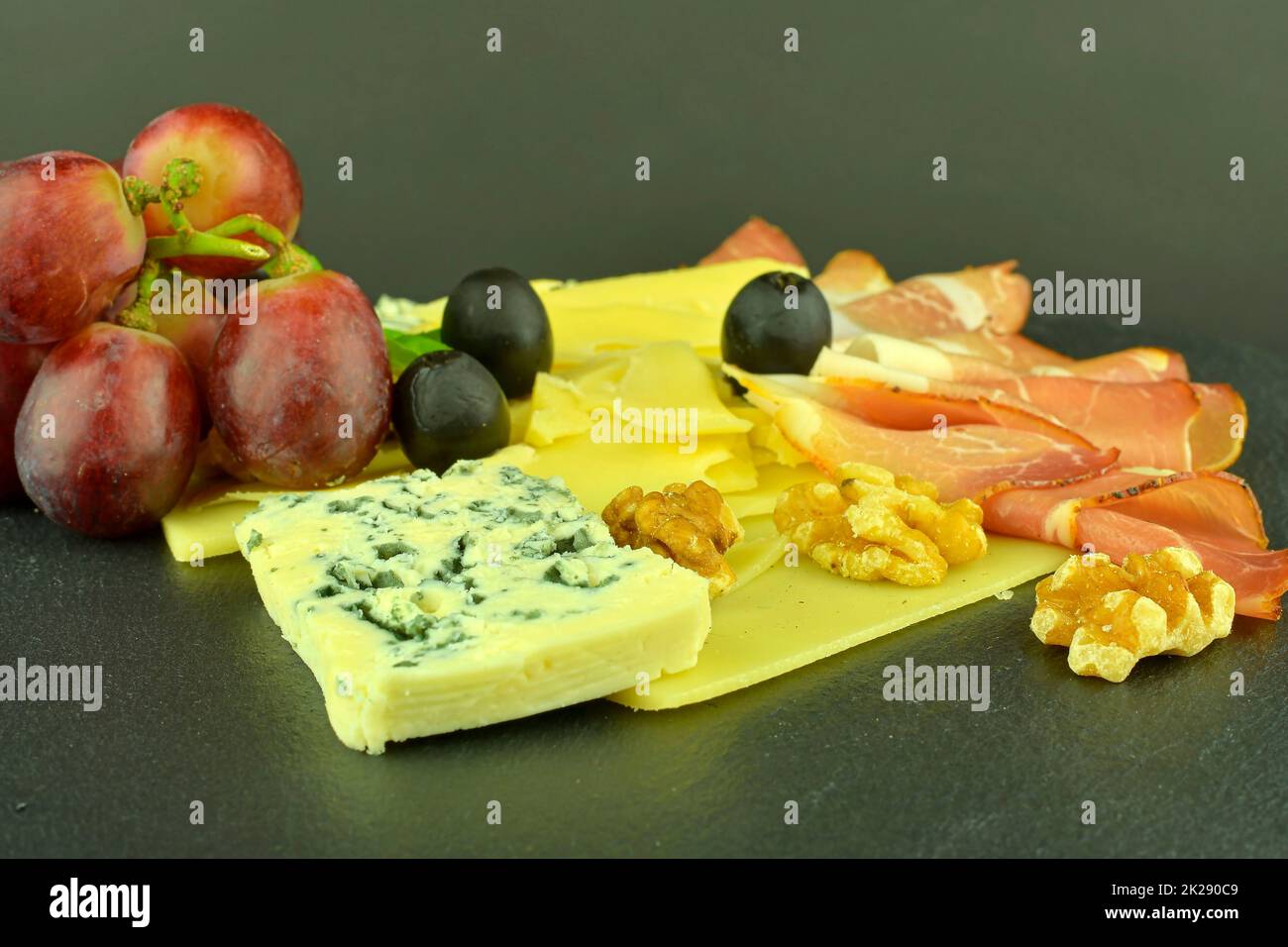 Assorted cheeses on black slate board. Brie cheese, cheddar slices, walnuts, grapes, olives and