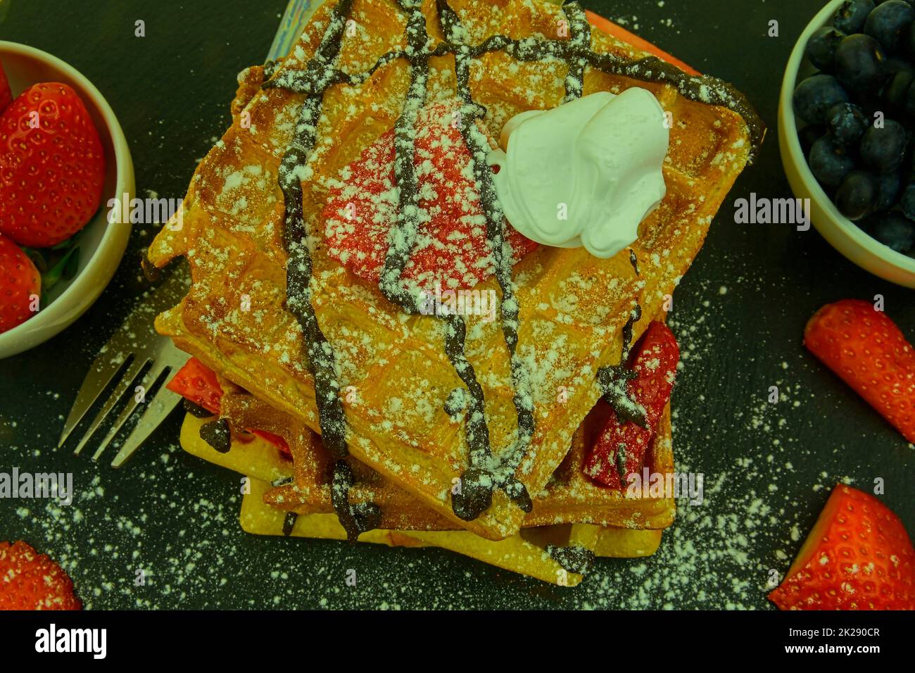 Square Belgian waffles with berries and icing sugar on a black plate. Tasty sweet sugary waffles. Melted chocolate and whipped cream on waffles. Top view Stock Photo