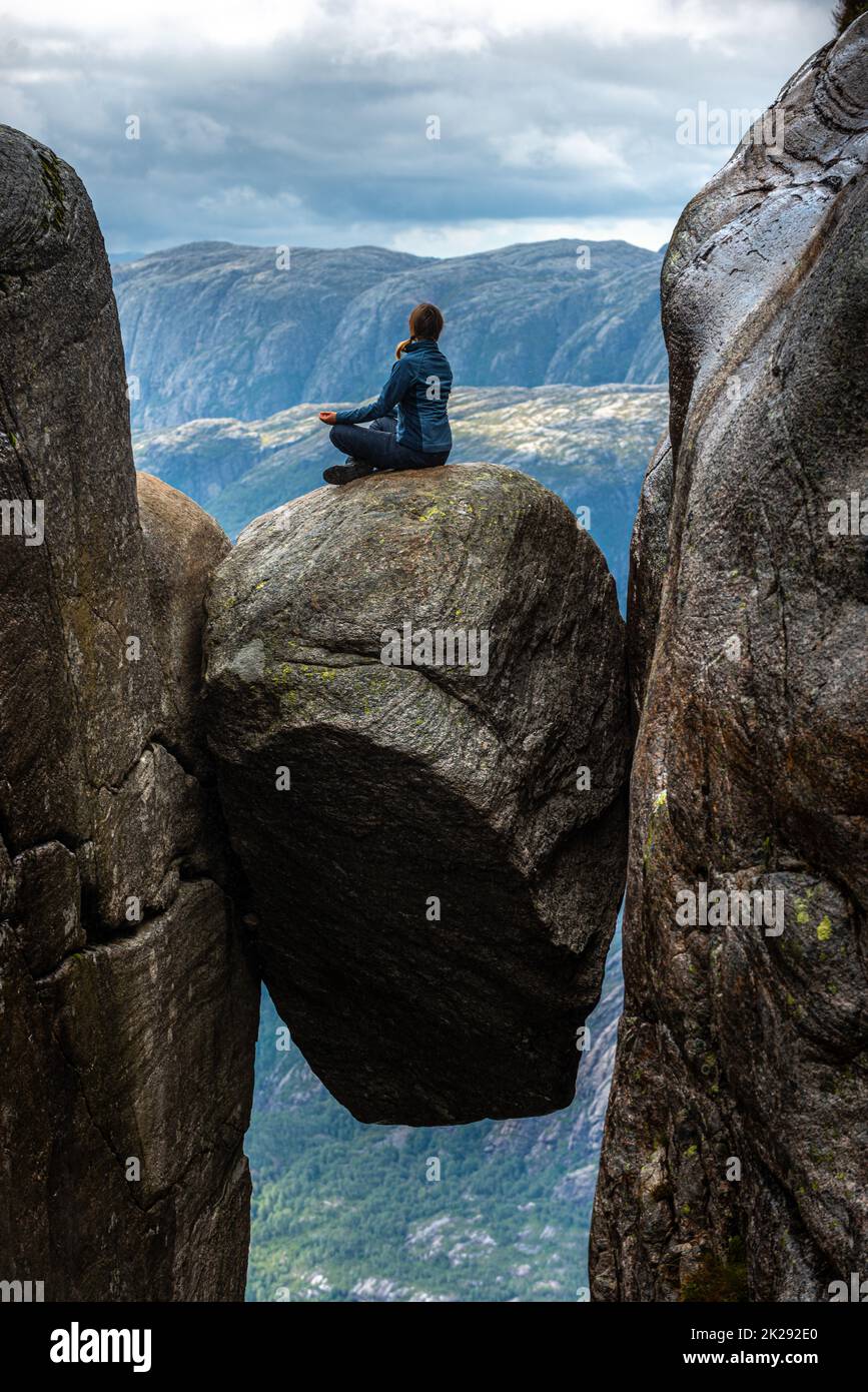 Visiting Norway Kjeragbolten located south of Lysefjorden Young woman sits on top of gian boulder in meditative pose. Stock Photo