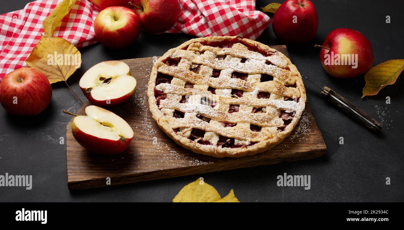 baked round traditional apple pie on brown wooden board and fresh red apples, top view Stock Photo