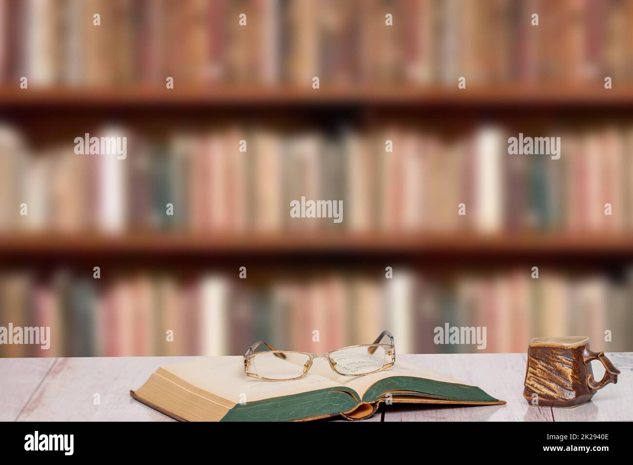 Books backgrounds. Closeup of an antique book with reading glasses on it and a brown clay mug on a light pink desk over a blurred bookshelf in library. Education concept. Teachers day. Stock Photo