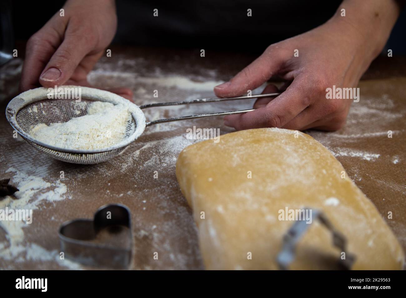 The chef's hands hold a sieve with flour on the background of parchment, flour, dough and cookie molds Stock Photo