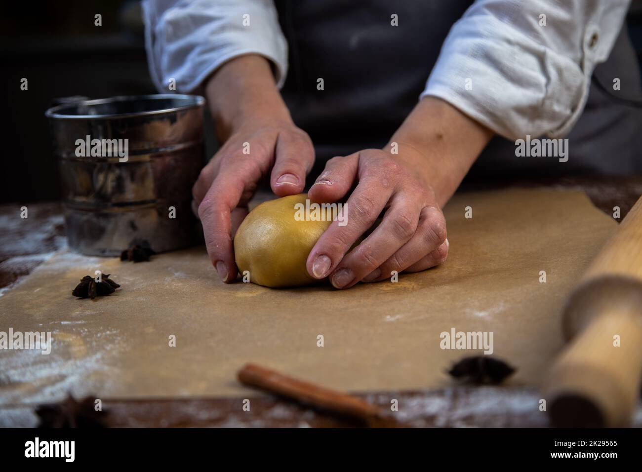 There is a dough on the parchment, slightly sprinkled with flour, which the cook holds in his hands, a rolling pin, cookie cutters, cinnamon and a metal flour jar are visible around. Stock Photo