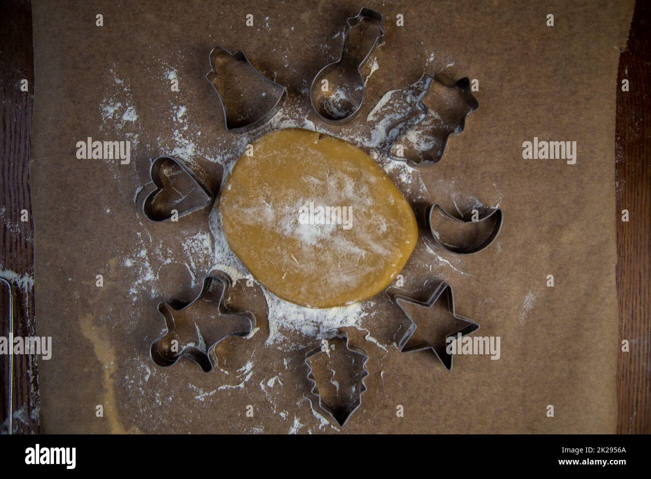 There is a dough on the parchment, around which there are cookie cutters of different shapes, all sprinkled with flour Stock Photo