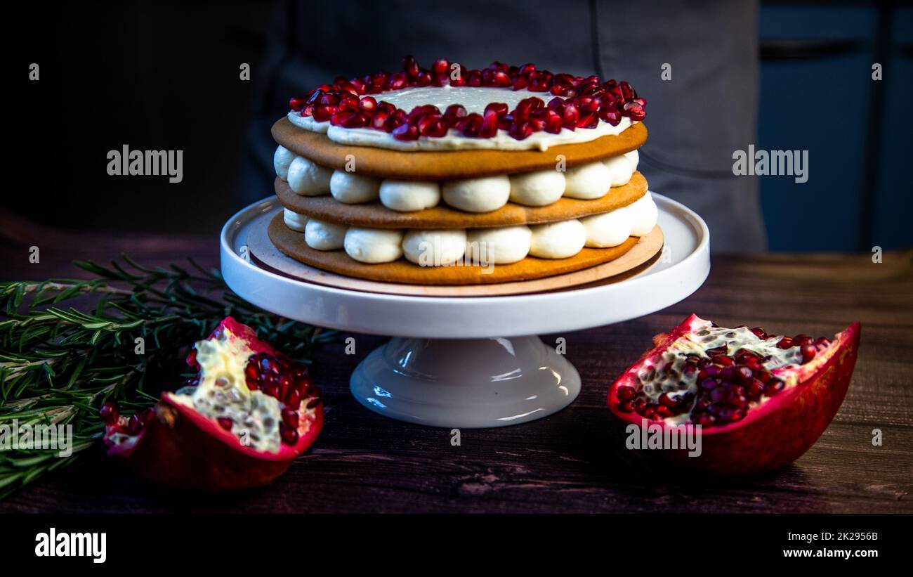 A cake of three layers, consisting of cakes and white cream, decorated with pomegranate and rosemary on top, stands on a white stand sideways on a dark background Stock Photo