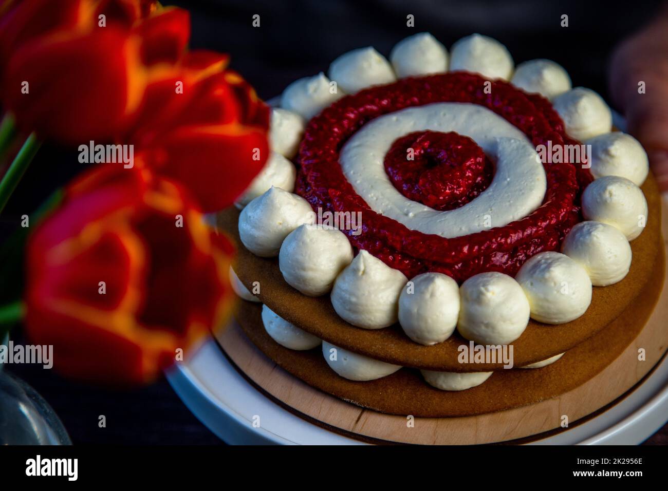 The baked cake cake lies on a wooden substrate and on a white stand, the cake is decorated with white cream and raspberry jam, in a circle, circles alternate, a bouquet of red tulips on a dark background is visible nearby Stock Photo
