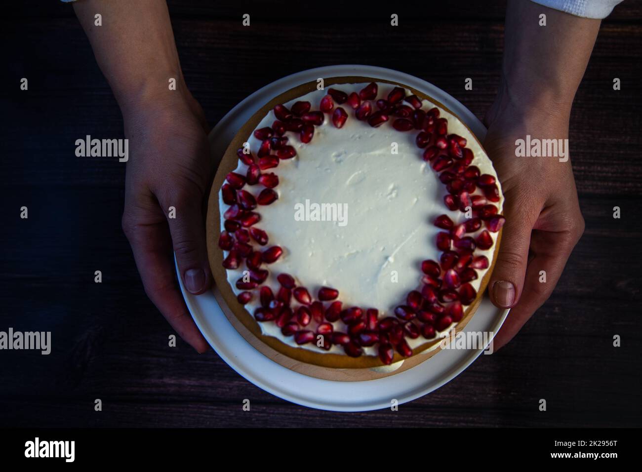 A cake of three layers, consisting of cakes and white cream, decorated with pomegranate on top, is held in the hands of a cook on a dark background, top view Stock Photo