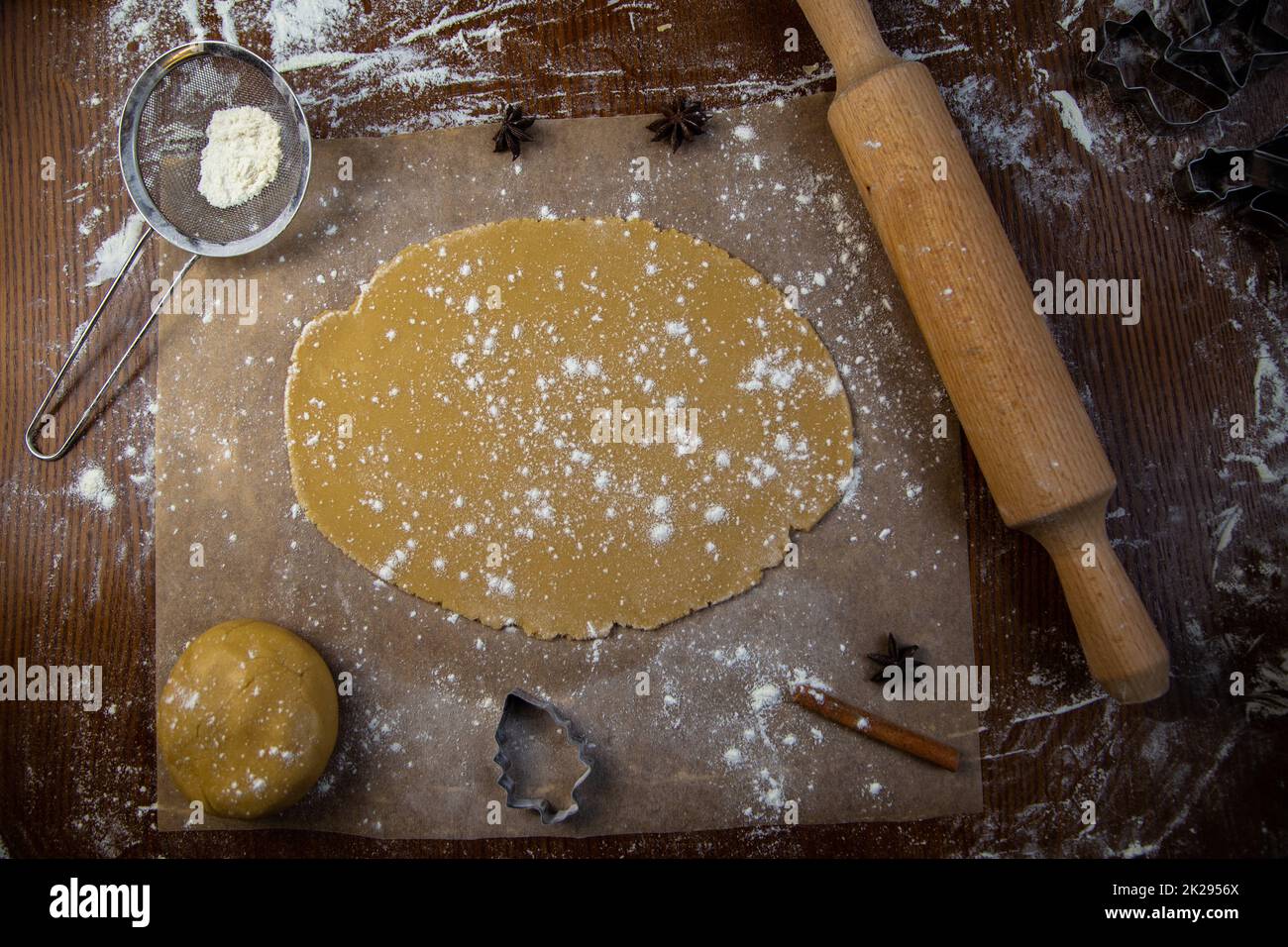 There is a rolled dough on the parchment, next to it is a lump of dough, a rolling pin, cookie cutters, cinnamon, everything is strewn with flour on a dark background Stock Photo